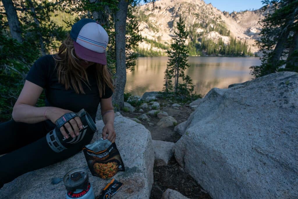 Kristen Bor pouring water from a Jetboil into a backpacking meal pouch next to a mountain lake