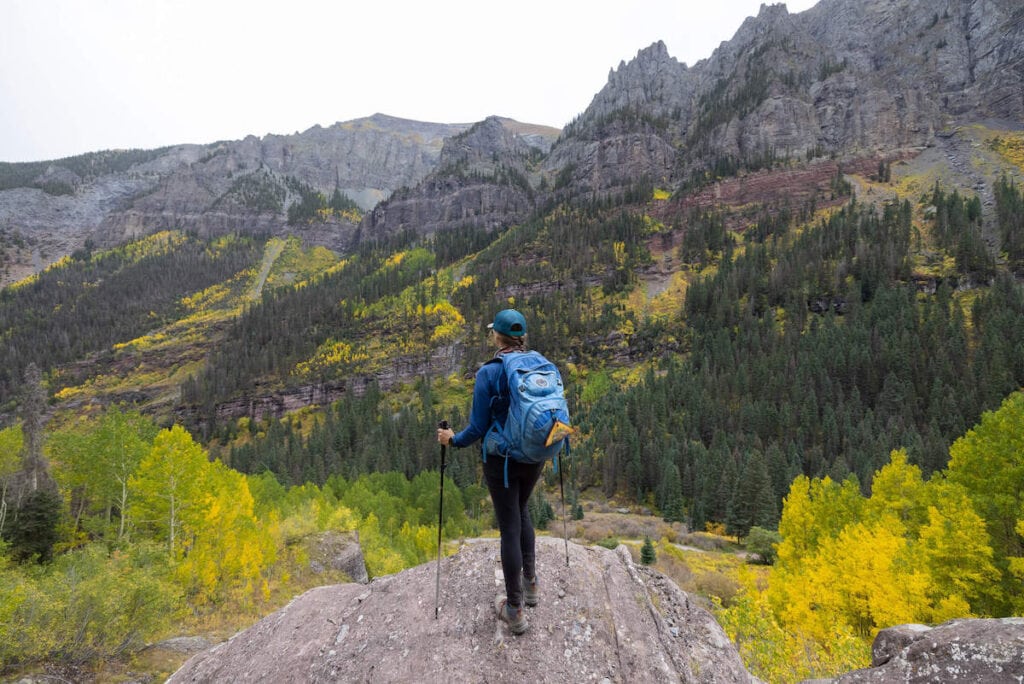 Woman standing on a rock wearing a day hiking pack looking out at the fall colors along the mountains