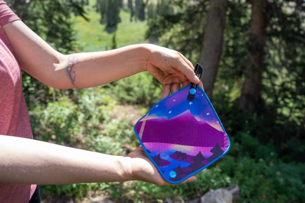 Woman holding a Kula Cloth pee rag featuring a bear next to a lake with a cosmic mountain scene