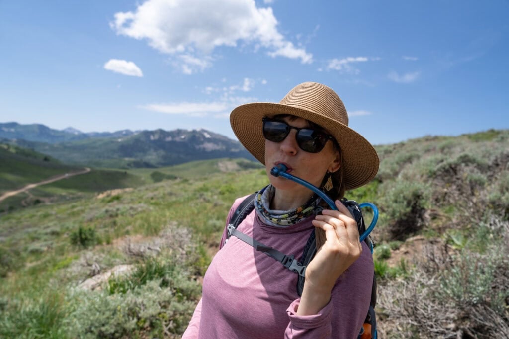 Kristen Bor on a hike with a water hose from a hydration reservoir in her mouth as she takes a drink of water