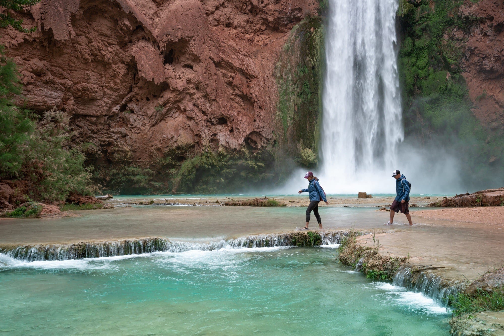 Two people walking in the turqoise water in front of Mooney Falls