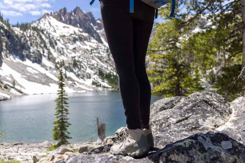 Photo of women's legs and high-ankle Oboz Bridger B-Dry Hiking Boot standing on rock overlooking alpine lake with snow-covered peaks in the background